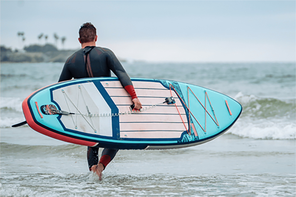 Brandon with paddleboard walking into the sea Brandon carrying a paddleboard at the sea